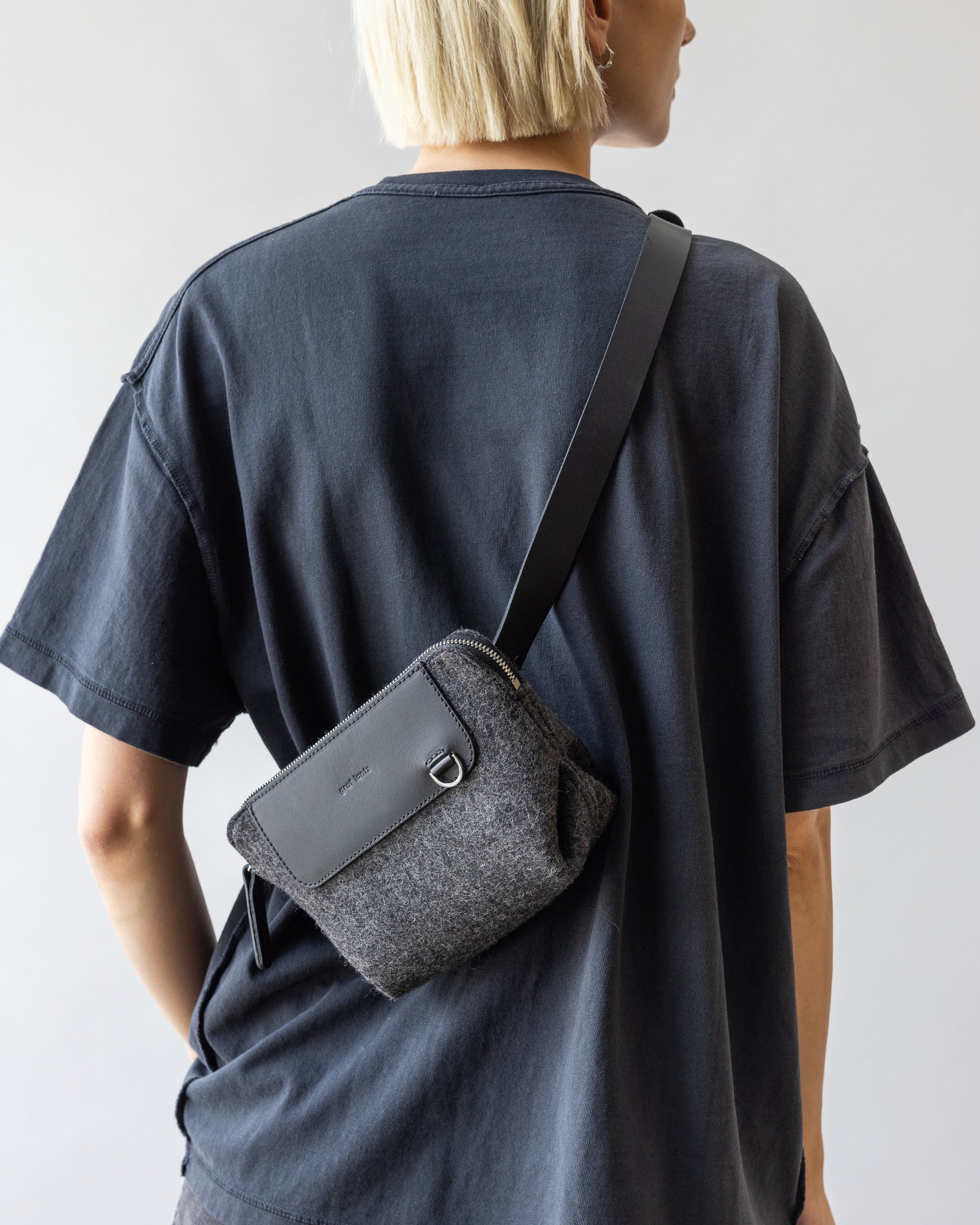 A person with short blonde hair, wearing a dark oversized T-shirt, sports a sleek graf lantz belt bag made of sustainable materials. The small dark gray bag features a black strap worn across their shoulder, contrasted against a plain light-colored background.