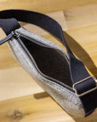 A gray graf lantz Merino Wool sling bag featuring a black adjustable strap and a partially open zipper sits on a light wooden surface, casting a soft shadow—ideal for holding your daily essentials.