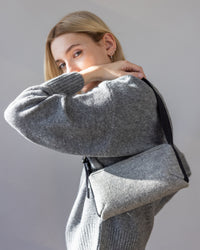A person with blonde hair wears a gray Merino wool sweater and holds a sleek graf lantz Campus Shoulder Bag. They pose confidently with an arm bent, gazing at the camera against a neutral backdrop.