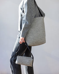 A person in a gray sweater and jeans carries a large gray graf lantz Campus Shoulder Bag over their shoulder and a small matching handbag in hand, ideal for everyday essentials. The scene is set against a plain, light background.