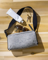 A graf lantz Campus Shoulder Bag with a black strap lies on a wooden surface. Next to it are sunglasses, a cream tube, keys on a ring, and a carabiner clip. The Merino Wool bag is slightly open and positioned at an angle.