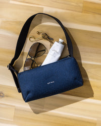 A minimalist blue graf lantz Campus Shoulder Bag with a sleek black strap holds sunglasses, keys with a gold hook, and a lotion tube. Set against a light wooden surface.