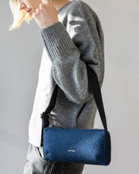 A person in a gray merino wool sweater and jeans carries a minimalist graf lantz Campus Shoulder Bag, showcasing a blue design with a black strap and subtle small text.