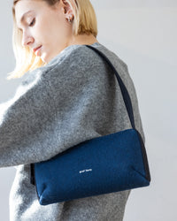 A short-haired person in a gray sweater admires a minimalist navy felt bag by graf lantz, crafted from Merino Wool, adding sophistication as they stand in a softly lit backdrop.