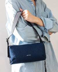 A person wearing a light blue denim shirt holds a graf lantz Campus Shoulder Bag in dark blue with a textured finish. The minimalist bag features a black strap and a small white logo.