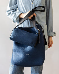 A person in a light denim shirt and jeans carries a minimalist dark blue felt handbag by graf lantz, featuring the words grote lente. The neutral background accentuates the simplicity of their campus shoulder bag.