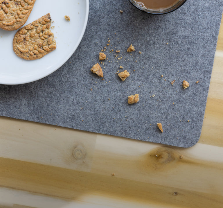 Top view of a wooden table with a grey felt placemat, a white plate of cookies, a drinking glass, and crumbs scattered on the placemat.