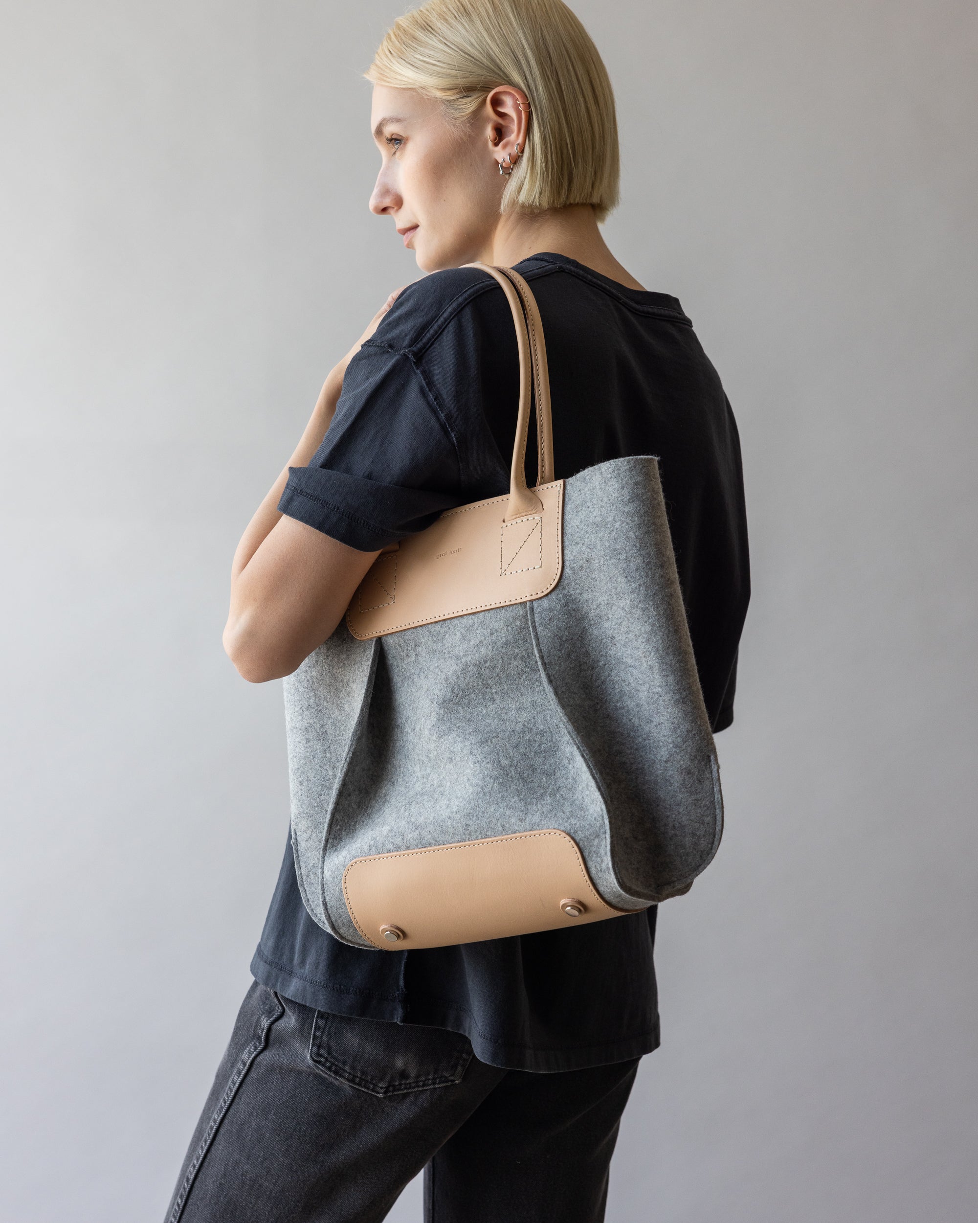 A person with short blonde hair is wearing a black T-shirt and holding a large gray and beige tote bag from graf lantz. This versatile accessory complements their style as they stand turned to the side against a plain backdrop.