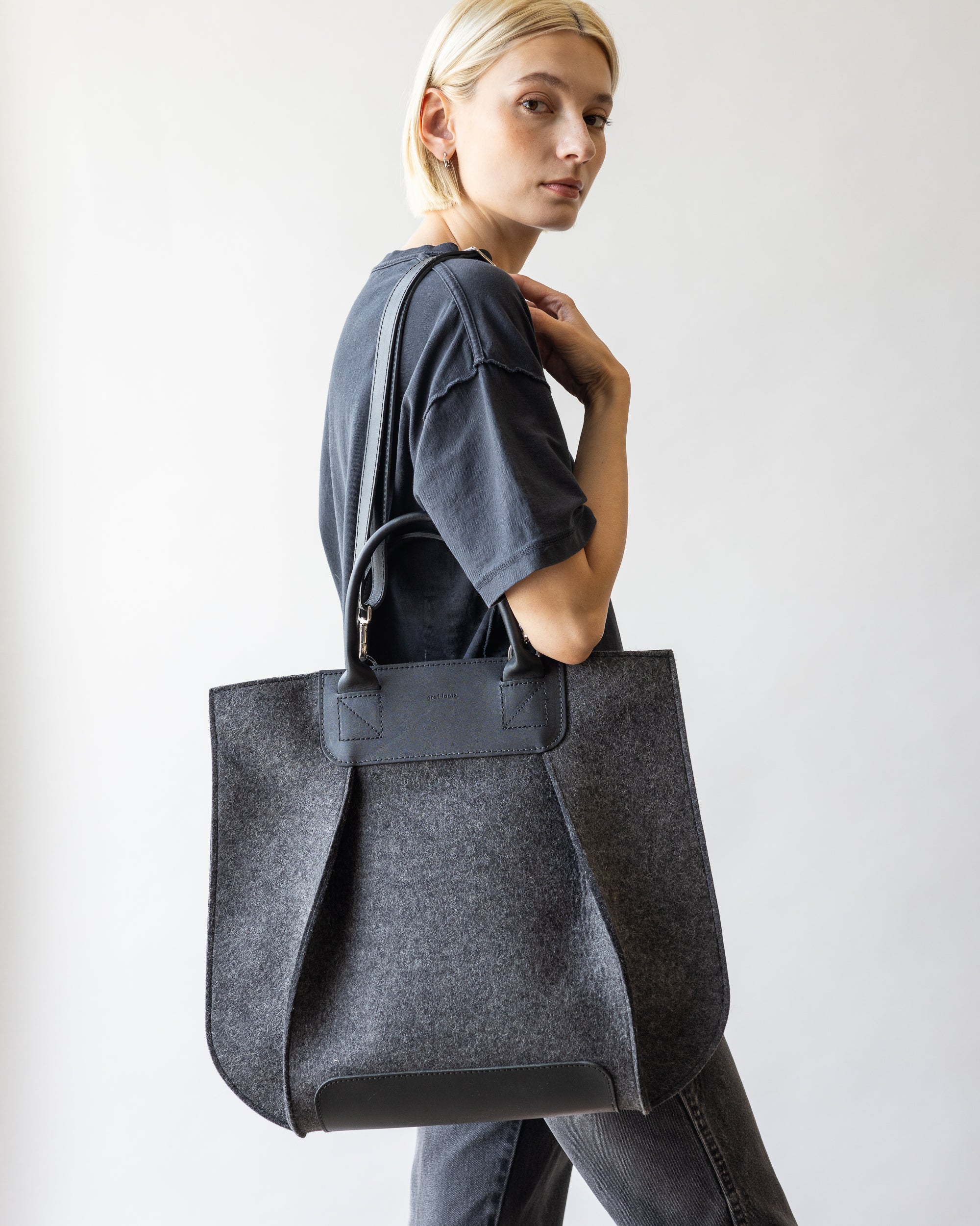 A confident individual with short blonde hair, wearing a dark t-shirt and jeans against a white background, carries the graf lantz Frankie Tote—featuring black handles and a structured design—in bold Merino wool over their shoulder.