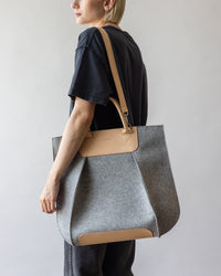 A person in black attire holds the versatile Frankie Tote by graf lantz—an elegantly structured gray bag with tan leather straps and accents. Set against a simple, light gray background, this tote blends style with functionality.