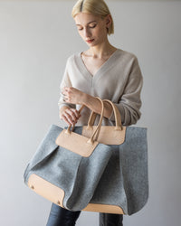 A person with short blond hair in a beige sweater holds the large Frankie Tote by graf lantz, featuring gray and tan hues with leather handles. The simple gray background adds a minimalist touch, enhancing the stylish look.