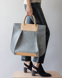 A person carries a large gray and beige Frankie Tote by graf lantz, crafted from Merino Wool, complementing their black leather pants and heeled ankle boots against a plain light gray backdrop.