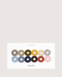 A set of 12 vibrant Wine-Os wine glass markers by graf lantz, made from Merino wool felt, includes colors like gray, green, olive, yellow, mustard, beige, blue, navy, teal, red, peach and pink. The box reads graf lantz and wine-Os.