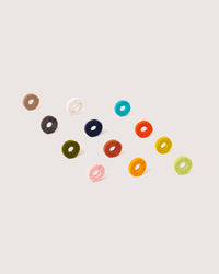 Twelve colorful, round felt rings from graf lantz are elegantly arranged on a light background as wine glass markers. With shades of brown, pink, blue, green, yellow, and orange featuring white centers, these sustainable luxury items add a chic touch to your gatherings.