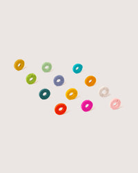 Graf Lantzs Wine-Os, small doughnut-shaped wine glass markers, are creatively displayed diagonally on a white background. Made from sustainable wool, they come in vibrant colors like yellow, green, blue, orange, purple, and pink.