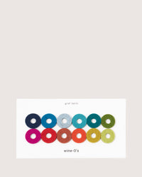 A white card displays 12 colorful wine-Os felt rings in two rows, made from Bavarian Merino wool by graf lantz. Each vibrant ring marks a wine glass in shades of blue, green, red, and pink.