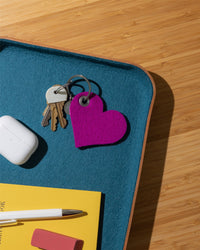 Graf Lantz Merino wool key fob and a bright pink heart-shaped keychain rest on a blue tray, alongside wireless earbuds, a yellow notebook, white pen, and pink eraser on a light wood surface.