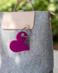 The Graf Lantz backpack features light gray felt, a tan leather flap, a magenta heart-shaped keychain, and a small round metal tag on the handle. Shown against blurred greenery, its a stylish, sustainable accessory.