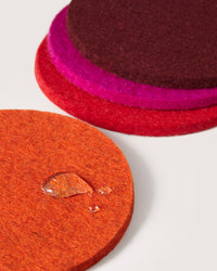 Stacked round coasters in shades of orange, red, pink, and burgundy crafted by graf lantz from sustainable German Merino wool. The close-up of the orange coaster reveals a water droplet on its surface against a neutral light background.