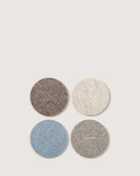 Four round felt coasters from graf lantz, made with premium German Merino wool, are arranged in a square on a plain background. They come in shades of brown, ivory, blue, and gray. The gray coaster displays graf lantz in white text.