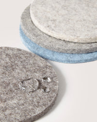 A stack of graf lantz round felt coasters in gray, blue, and cream rests on a light surface. Made from German Merino wool, the top coaster sparkles with droplets, showcasing its soft, fuzzy texture.