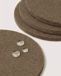 Close-up of round brown Graf Lantz Merino wool coasters on a white surface, with three water droplets beading on the front coaster, highlighting its water-resistant feature—a stylish, sustainable home decor choice.