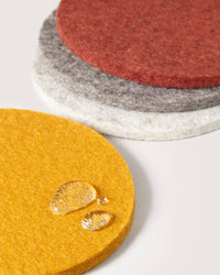 A set of graf lantz round Merino wool coasters in yellow, white, gray, and red is showcased. The foreground features a yellow coaster with water droplets, highlighting its water resistance. Perfect as beer felt coasters, they are placed on a plain light-colored surface.