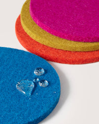 Graf Lantz presents stacked Merino wool felt coasters in vibrant blue, orange, yellow, and pink. The water-resistant surface of the blue beer felt coaster showcases how effortlessly water droplets bead on it.