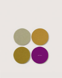 Four round Graf Lantz felt coasters in green, mustard yellow, ochre, and purple German Merino wool are arranged in a square on a white background. The purple coaster displays the Graf Lantz logo in small white letters.