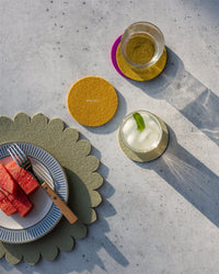 A plate of watermelon and a fork rest on a green scalloped placemat, with drinks on colorful Graf Lantz felt coasters made from German Merino wool atop a light gray surface.