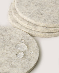 Close-up of stacked, round light gray felt coasters by graf lantz, made from soft Merino wool. One coaster in the foreground shows water droplets, highlighting its absorbent texture—ideal for keeping surfaces dry during a cold beer.
