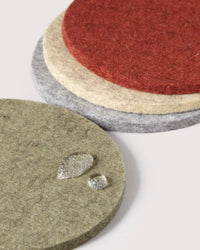 Four round coasters by graf lantz, crafted from German Merino wool, come in green, gray, beige, and red. They are stacked and slightly overlapping on a neutral background. The top green coaster features water droplets for an added touch of sustainable home décor elegance.