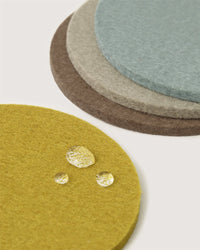 Four round Graf Lantz Merino wool coasters in yellow, brown, gray, and blue are stacked on a white background. The yellow coaster is in front with three water droplets. Made from German Merino wool felt.