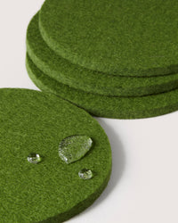 Graf lantz green felt coasters, crafted from premium German Merino wool, are artfully stacked and scattered. Three water droplets bead on the foreground coasters surface, highlighting its water-resistant texture against a crisp white background.