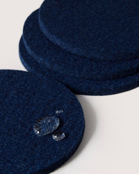 A stack of round dark blue felt coasters by graf lantz, crafted from premium Merino wool, is displayed. One coaster stands out with water droplets showcasing its excellent water resistance, set against a neutral off-white background.