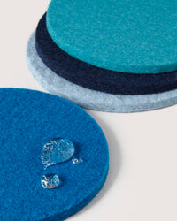Four round coasters from graf lantz, crafted from premium German Merino wool in various shades of blue, are stacked with one placed separately showing water droplets that highlight its excellent absorption. The background is plain off-white.