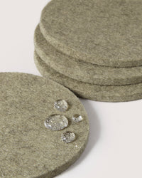 Close-up of stacked, round graf lantz felt coasters in a greyish-green hue made from Oeko-Tex Certified Merino wool. Water droplets glisten on the coaster in the foreground, showcasing its water-resistant surface against a plain white background.