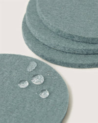 A close-up of teal Merino wool coasters by Graf Lantz, with one in the foreground featuring water droplets—an elegant and sustainable home decor choice.