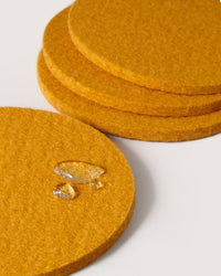 A close-up of four mustard-colored coasters from graf lantz, crafted from fine German Merino wool, stacked and spread out. The nearest coaster shows water droplets on its textured surface against a plain white background.