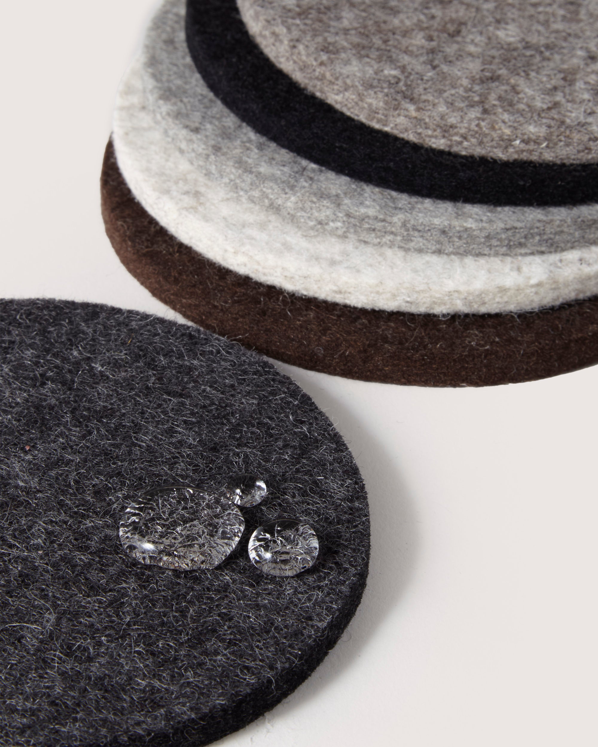 A stack of graf lantz circular Merino wool coasters in brown, gray, black, and white sits on a white surface. Water droplets highlight the texture on the foreground gray coaster made of German Merino wool.