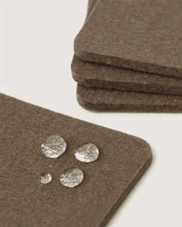 Close-up of brown Merino wool coasters by Graf Lantz, showcasing water droplets beading on their water-resistant surface. Three are neatly stacked in the background—a stylish, sustainable home décor accent for any space.