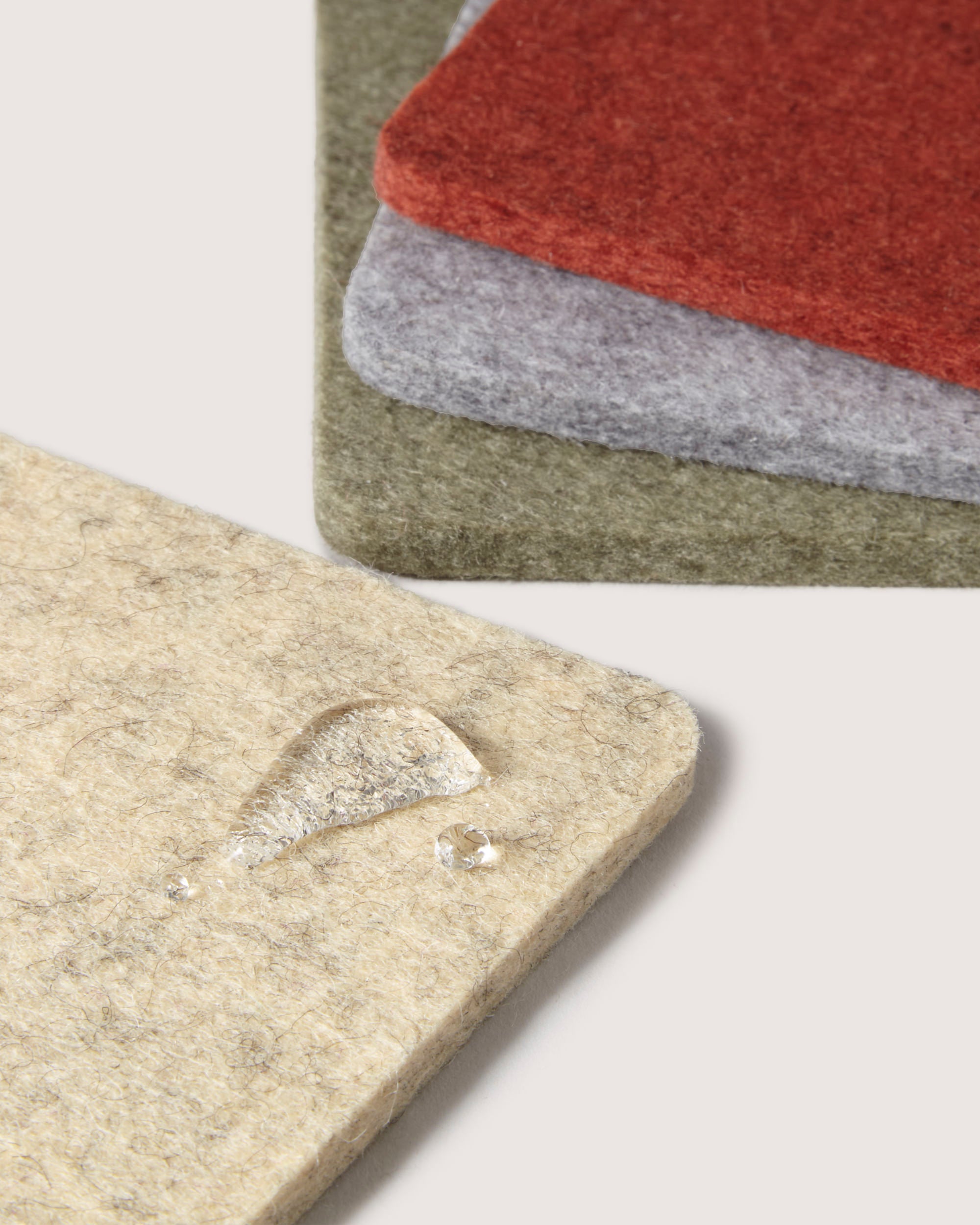A close-up shows four graf lantz Merino wool coasters in beige, olive green, gray, and red, stacked slightly off-center. A water droplet on the beige coaster highlights its texture and water resistance, perfect for your favorite beer mug or glass.
