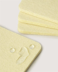 Close-up of off-white Bierfilzl Merino Wool Square Coasters, with three stacked behind. The top coaster has three light-reflecting water droplets, showcasing the soft texture of its felt surface..