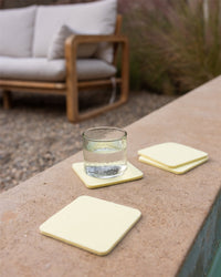 A glass of water sits on a light yellow Bierfilzl Merino Wool Square Coaster, placed on a stone ledge. Two additional coasters are nearby, while a wooden-framed couch with white cushions rests on the gravel surface in the background.