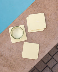 A glass with a pale yellow drink sits on a textured beige surface by a pool. It rests on the Bierfilzl Merino Wool Square Coaster from the Solid 4 Pack in light yellow, while another coaster is nearby. The blue water and paving stones are partially visible.
