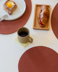 A breakfast setup shows a cup of coffee on a Bierfilzl Merino Wool square coaster, a fruit-filled puff pastry on a white plate, and an icing sugar-dusted croissant. These items sit on brown circular placemats.