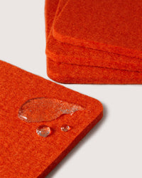 Close-up of graf lantz red felt coasters made from premium Merino wool with rounded edges. One coaster sparkles with water droplets, showcasing its water-resistant nature. The coasters are stacked elegantly against a plain gray background.