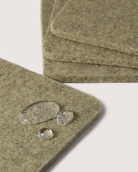 Close-up of stacked square coasters by graf lantz, made from textured, absorbent German Merino wool with soft beige tones. Water droplets enhance their natural appeal against a minimalist backdrop. Ideal for refined settings.