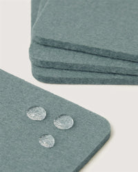Close-up of blue-gray Graf Lantz Merino wool coasters with three water droplets beading on the surface, showcasing water resistance. Additional sustainable German Merino wool coasters are stacked in the background on a white surface.