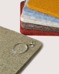 A stack of colorful graf lantz felt squares in red, orange, blue, and gray sits on a neutral surface. In the foreground is a beige Merino wool square with water droplets highlighting its texture and water resistance, ideal for stylish coasters.